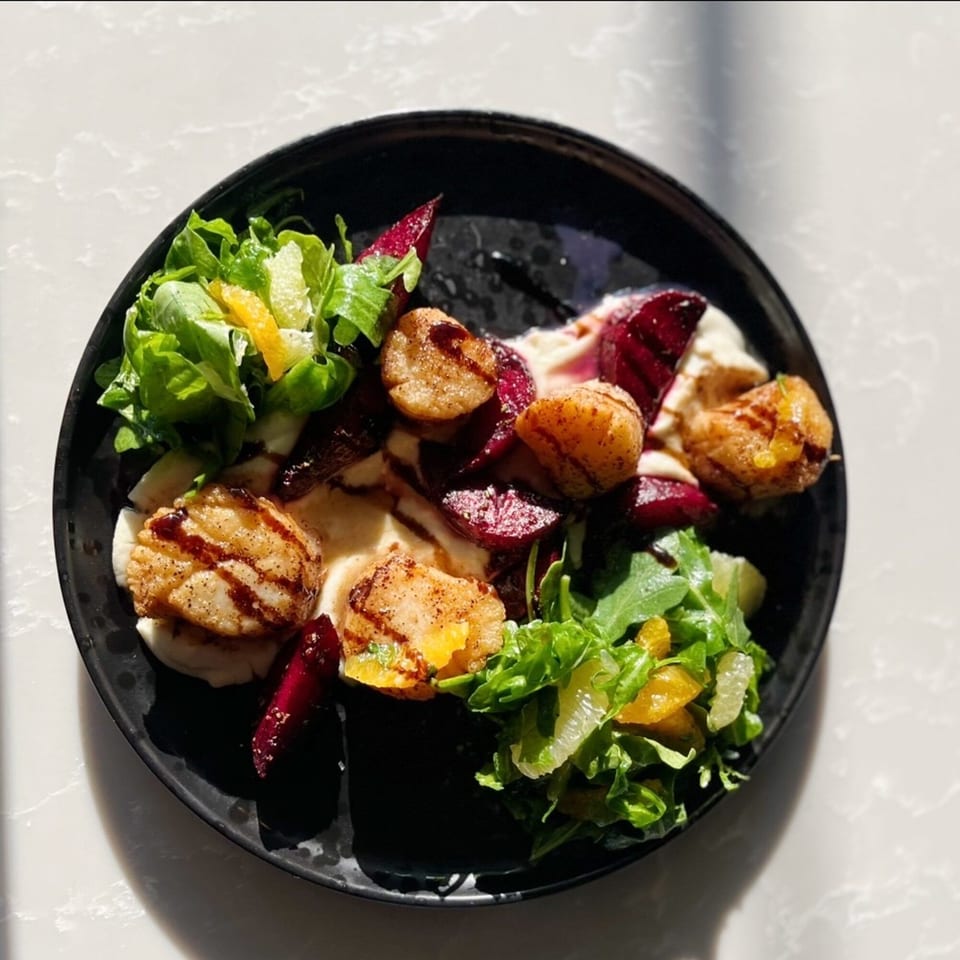 Seared scallops with beets, greens, and citrus segments on a black plate.