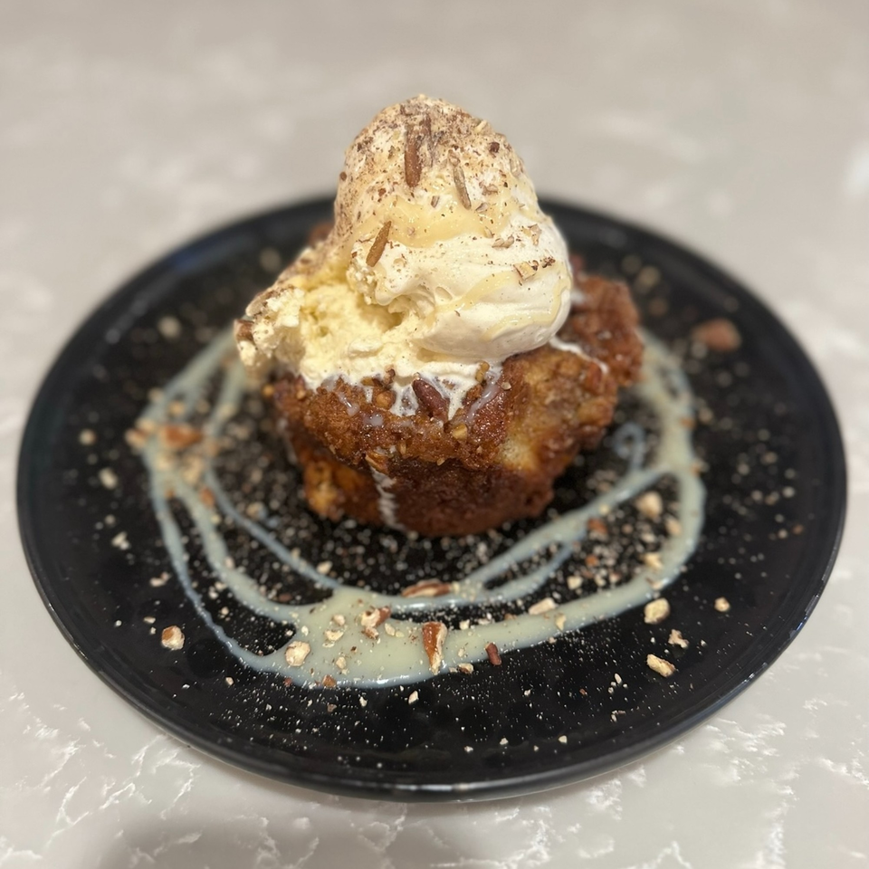 Warm bread pudding topped with vanilla ice cream, nuts, and a drizzle of white sauce on a black plate.