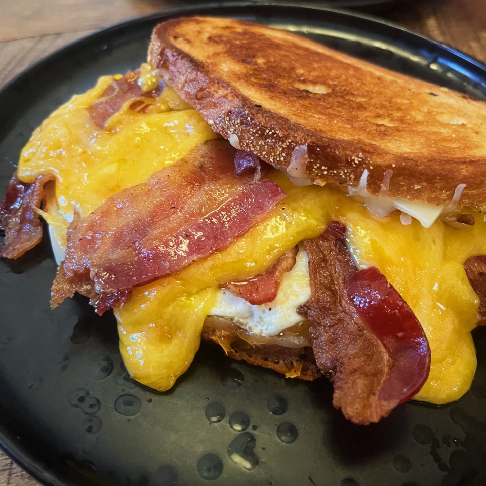 A grilled cheese sandwich with melted yellow cheese, bacon, and a fried egg on a black plate.