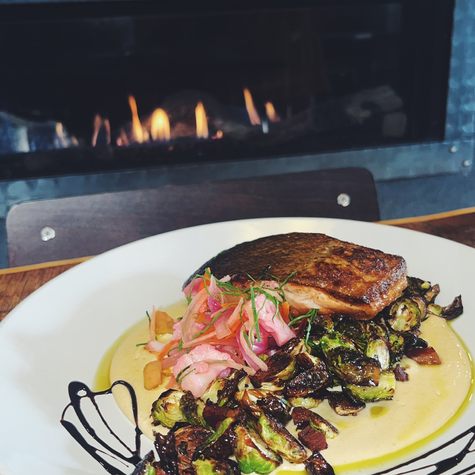 Seared salmon with roasted brussels sprouts and pickled vegetables on a white plate, with a fireplace in the background.